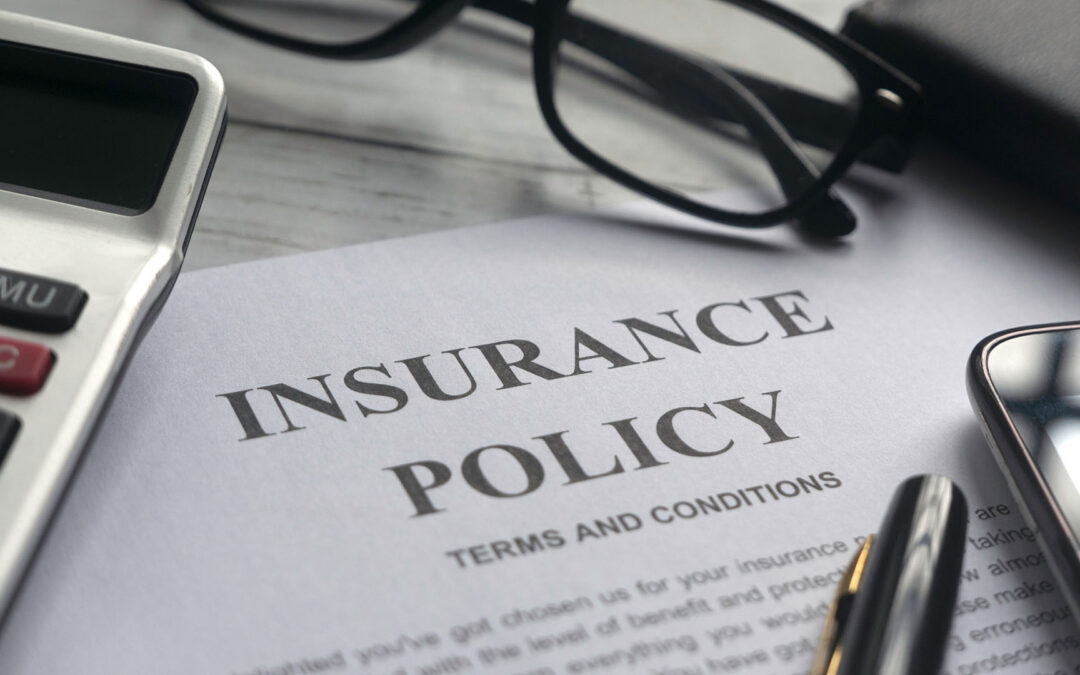 An insurance policy document titled "INSURANCE POLICY" in bold, capital letters, with "TERMS AND CONDITIONS" printed smaller below it. The paper is surrounded by a calculator, a pair of black-rimmed glasses, a pen, and a smartphone on a wooden surface. - https://www.staplesagency.com/