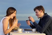 Man kneeling and holding an open ring box, proposing to a woman sitting across from him at an outdoor table