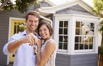 Smiling young couple holding house keys in front of a gray single-family home with white-trim windows.