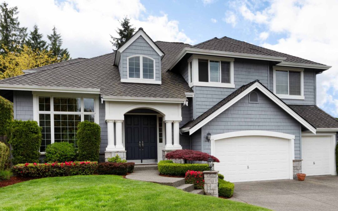 Spacious two-story suburban home with gray siding, attached two-car garage, manicured lawn, and front garden under a partly cloudy sky.
