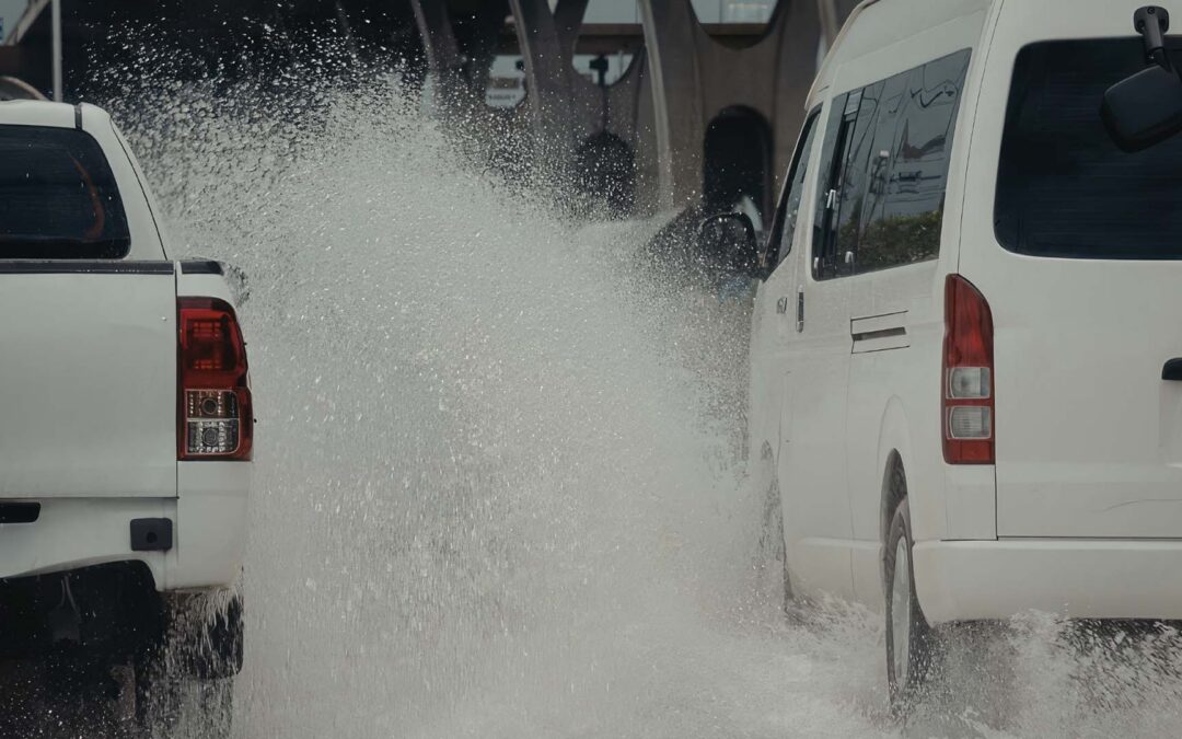 Two vehicles, a white pickup truck on the left and a white passenger van on the right, drive through a deep pool of water on a flooded road - https://www.staplesagency.com/
