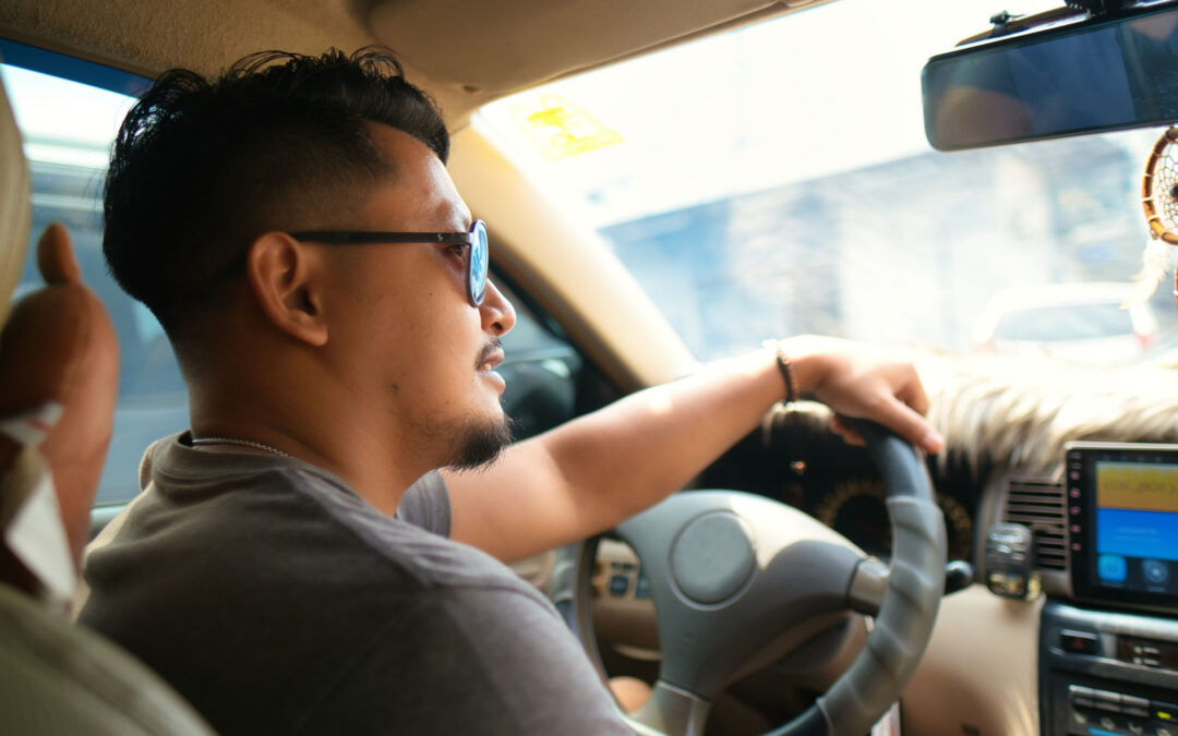 Driver at the wheel of a car, steering with one hand while a passenger gestures from the front seat; dashboard and hanging ornament visible.