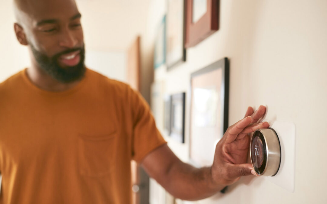 An African American man with a beard and a smile, wearing an orange t-shirt, is adjusting a round, silver smart thermostat on a white wall - https://www.staplesagency.com/