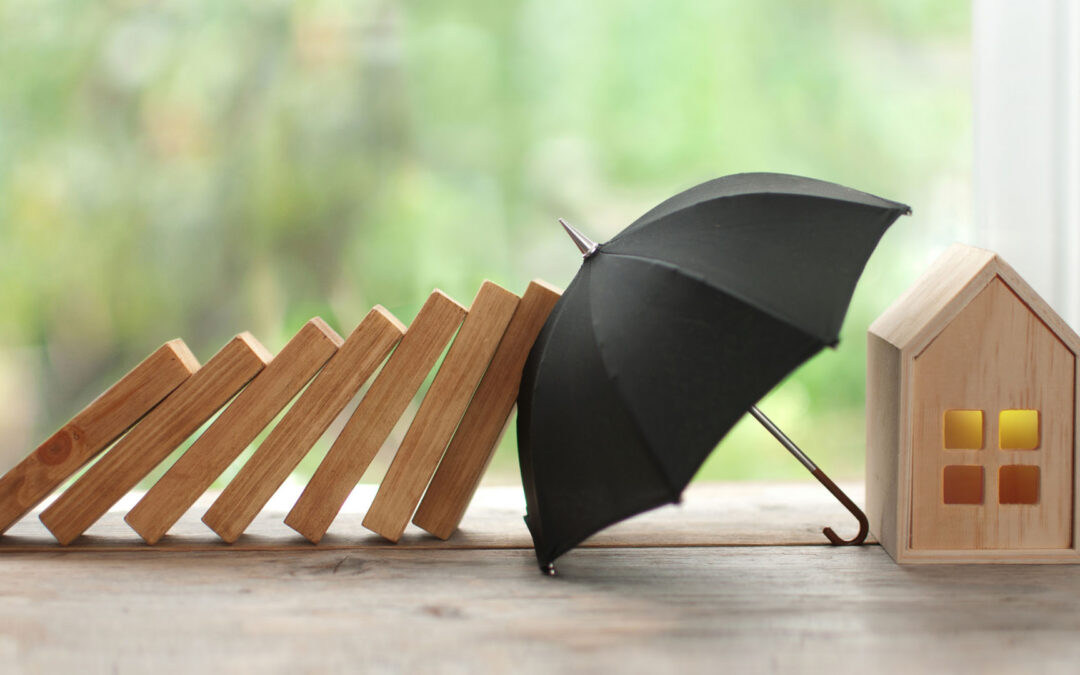 a small wooden house model protected by a black umbrella from a row of falling wooden blocks - https://www.staplesagency.com/
