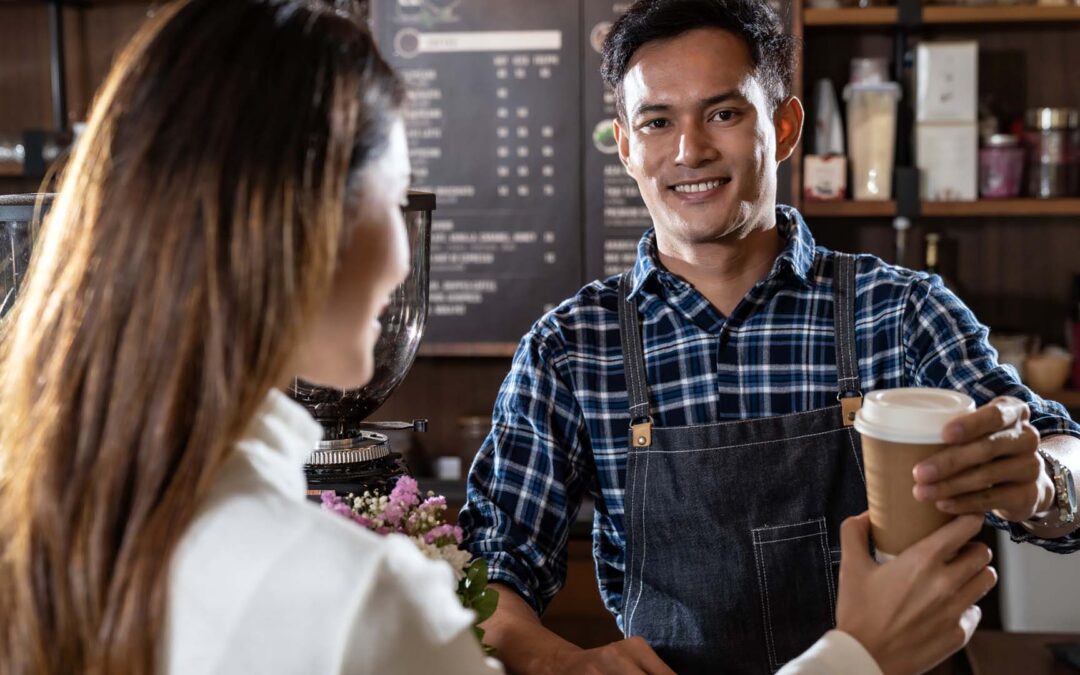 A smiling barista in a blue plaid shirt and dark apron hands a coffee in a paper cup to a customer with long brown hair in a cafe - https://www.staplesagency.com/