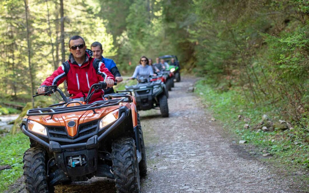 An orange all-terrain vehicle (ATV) leads a group of riders on a rocky path through a dense, sun-dappled forest - https://www.staplesagency.com/