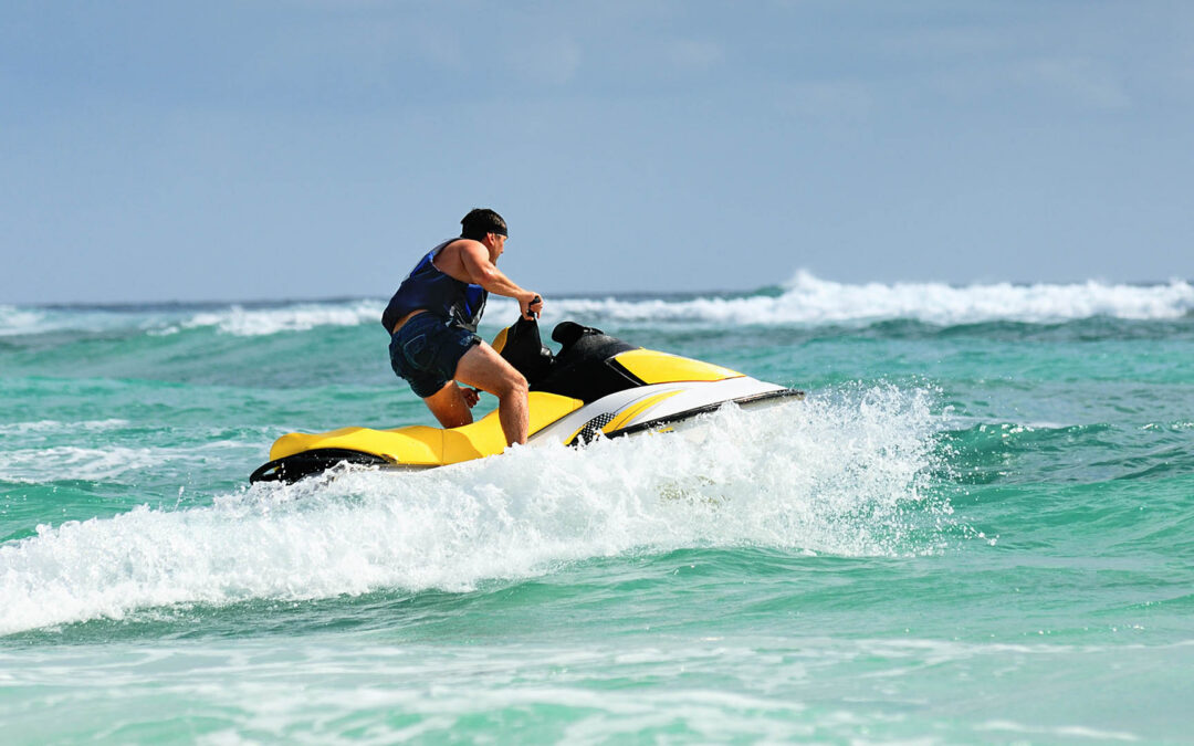 A man in a life vest rides a yellow and white jet ski through turquoise ocean waves, kicking up a large spray of water - https://www.staplesagency.com/