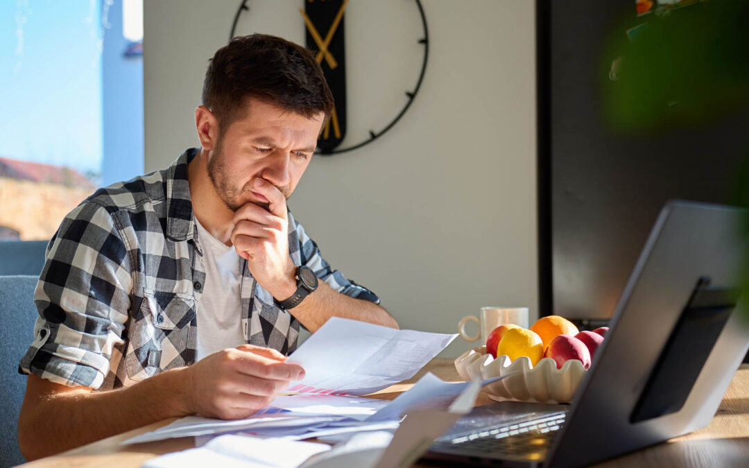 A man in a black and white checkered shirt sits at a sunlit wooden table, deeply focused on reviewing a stack of papers in his hands - https://www.staplesagency.com/