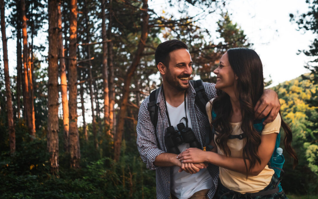 A happy couple hiking in a sunlit forest, with backpacks and a pair of binoculars - staplesagency.com