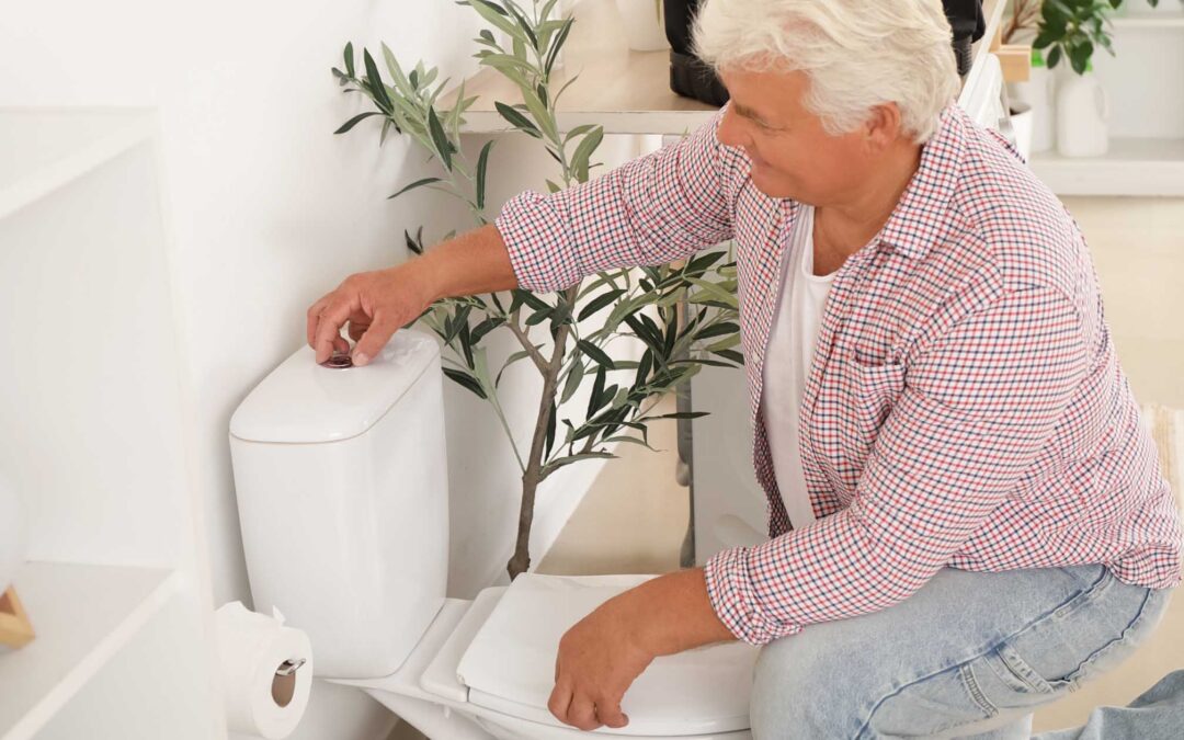 Elderly man in a checkered shirt crouches beside a white toilet - staplesagency.com