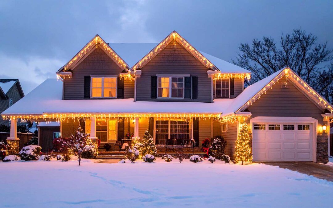 A two-story suburban home decorated with warm yellow icicle Christmas lights along its snowy roofline and porch at dusk - www.staplesagency.com