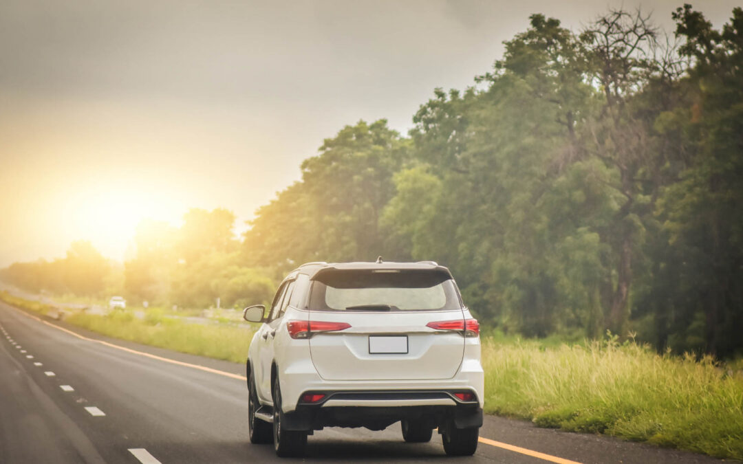 A white SUV travels down a straight highway bordered by green trees under a hazy, golden sun - www.staplesagency.com