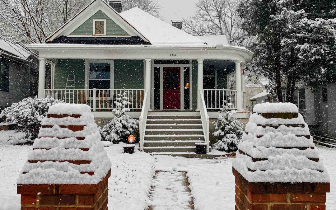 A snow-covered green bungalow with a red door and a white porch is framed by two snow-capped brick pillars in the foreground during a snowfall - www.staplesagency.com
