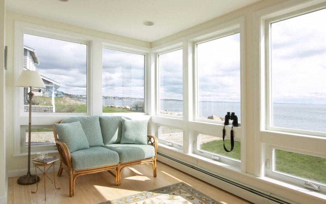 A bright sunroom features a light blue wicker loveseat positioned in front of large floor-to-ceiling windows that offer a panoramic view of a calm ocean coastline under a cloudy sky - www.staplesagency.com