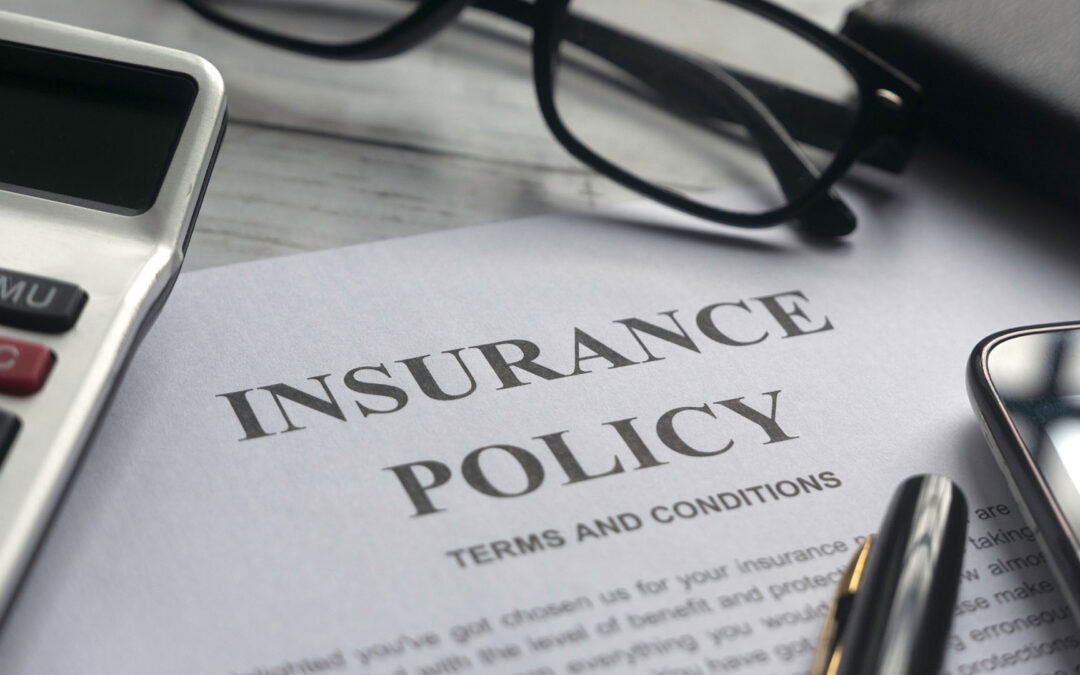 An insurance policy document titled "INSURANCE POLICY" sits on a desk surrounded by a calculator, a pair of eyeglasses, and a pen - www.staplesagency.com