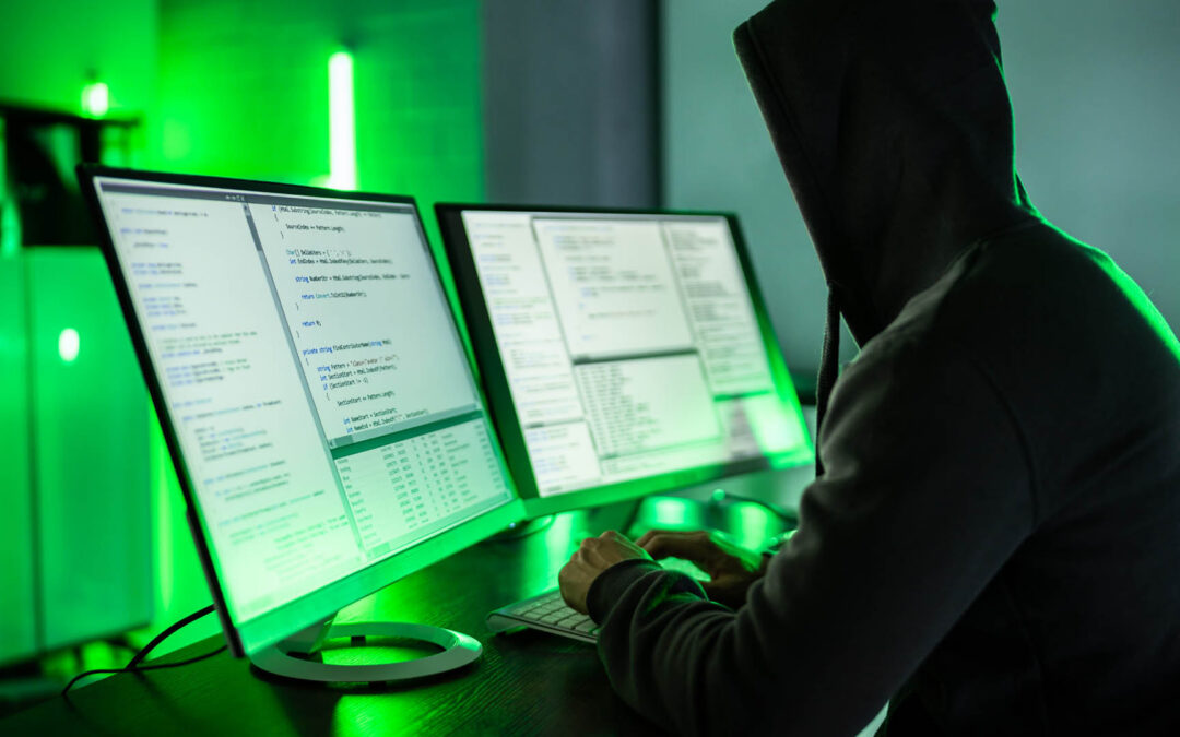 A hooded person sits in a dark room with green lighting, typing on a keyboard in front of two computer monitors displaying lines of code - www.staplesagency.com
