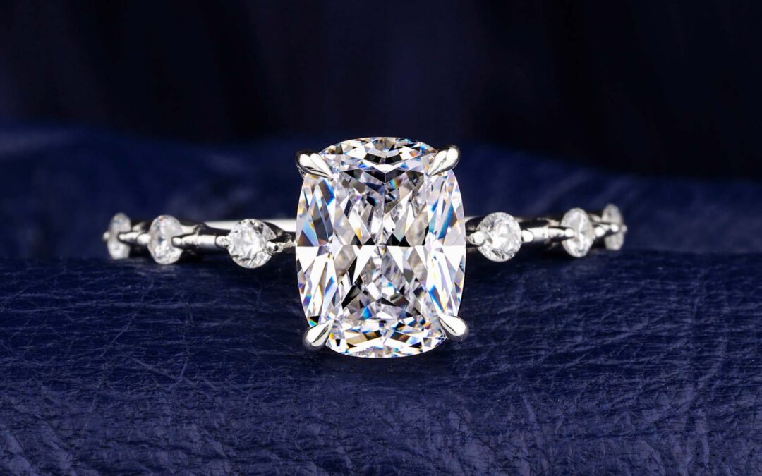 A cushion-cut diamond engagement ring with a delicate, diamond-accented band is centered against a textured dark blue background - www.staplesagency.com