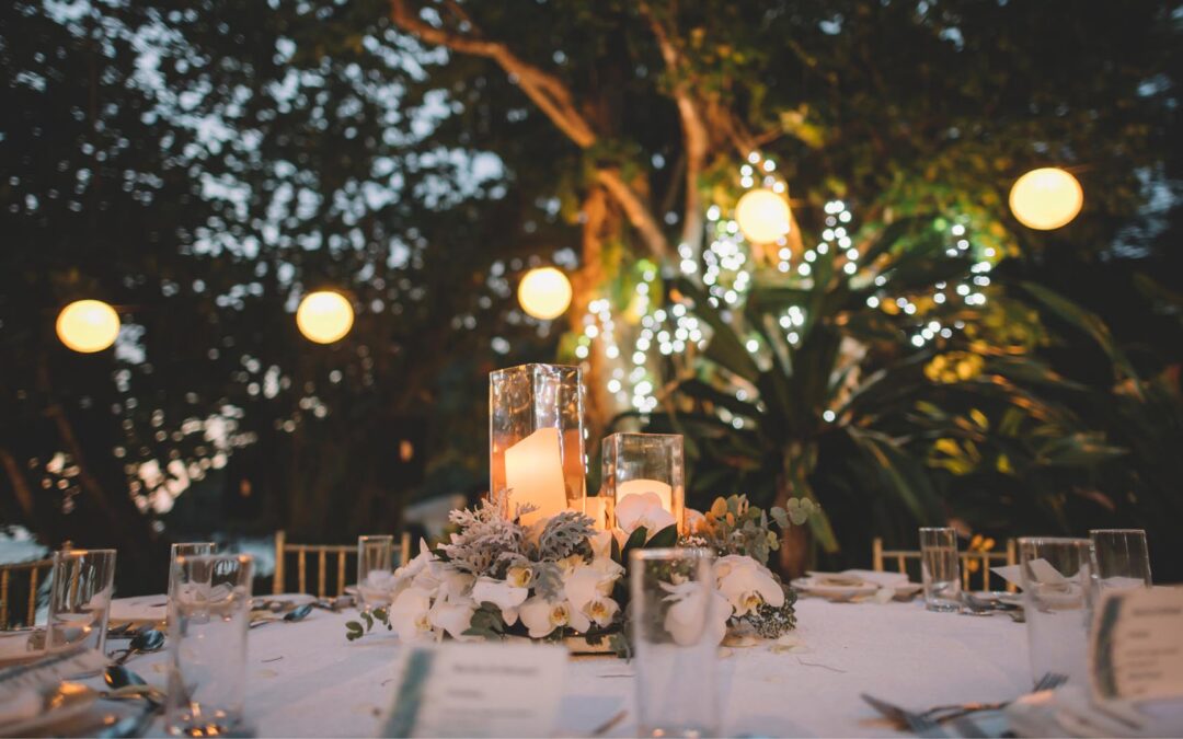 An outdoor evening wedding reception table is elegantly decorated with white orchids, dusty miller foliage, and glowing pillar candles in glass cylinders, set against a backdrop of twinkling fairy lights and hanging paper lanterns in the trees - www.staplesagency.com