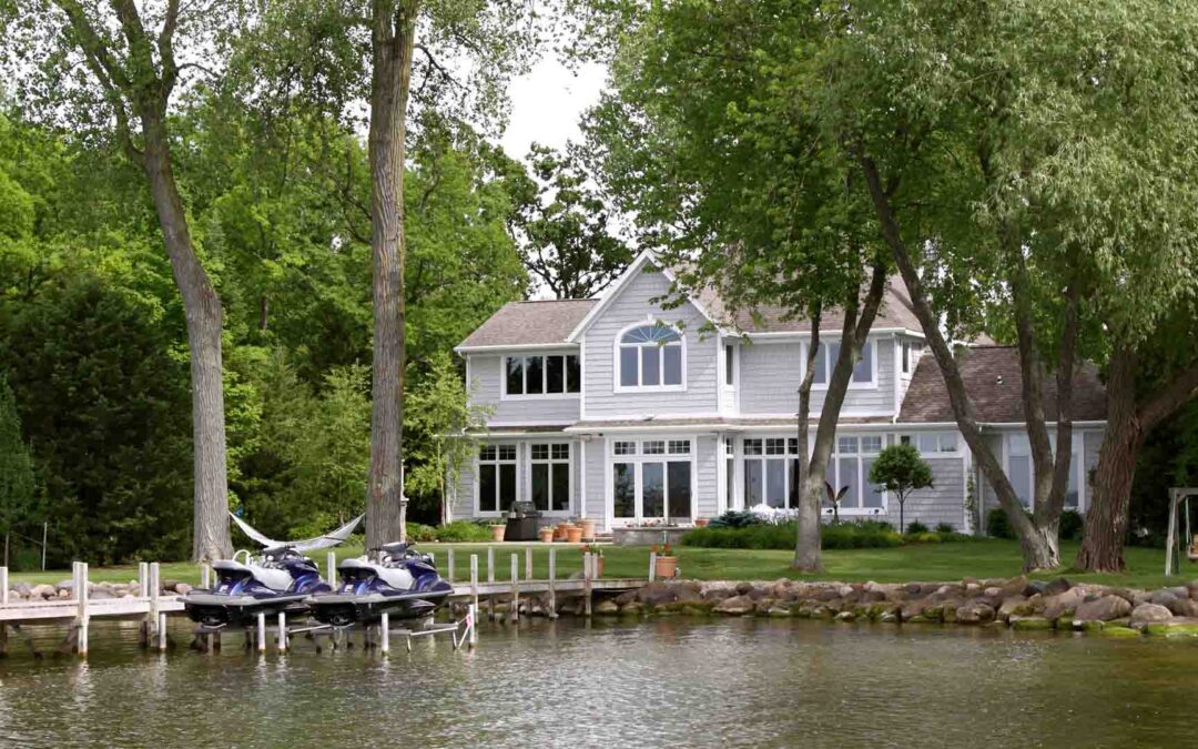 A large, two-story light gray lakefront home with many windows and a small dock featuring two jet skis sits nestled among lush green trees on a calm waterfront - www.staplesagency.com