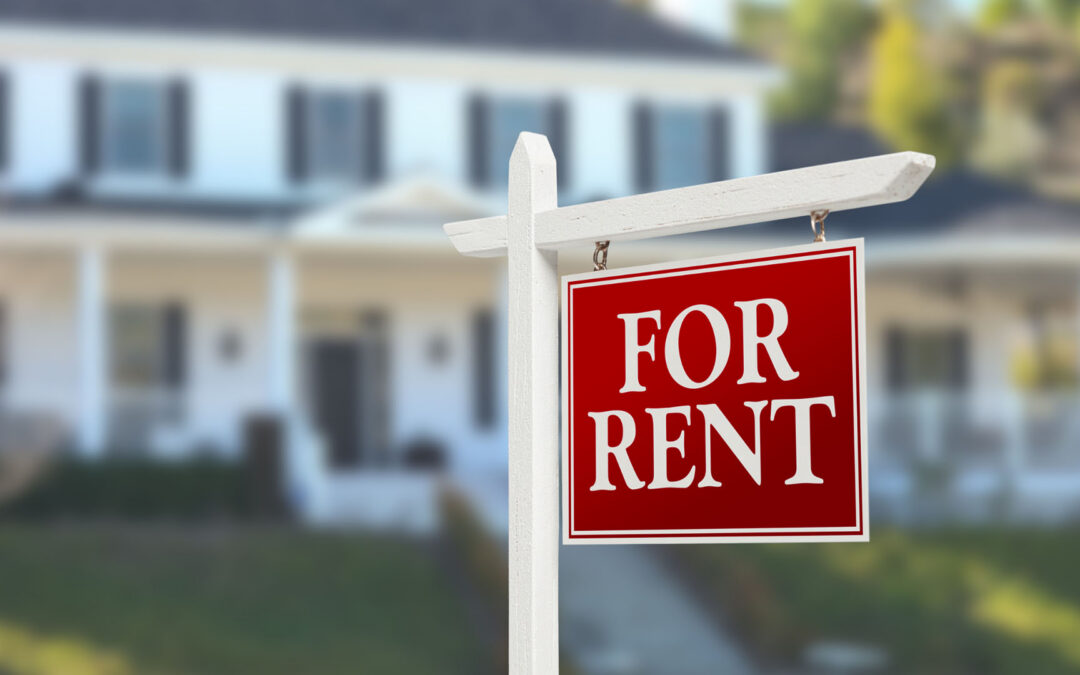 A red "For Rent" sign hangs from a white wooden post in front of a blurred, large white house - www.staplesagency.com
