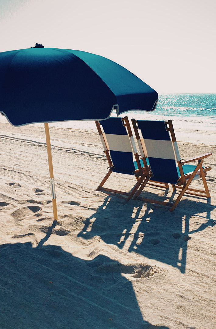 A pair of blue and white beach chairs under a blue beach umbrella sitting on a sandy beach facing the ocean.